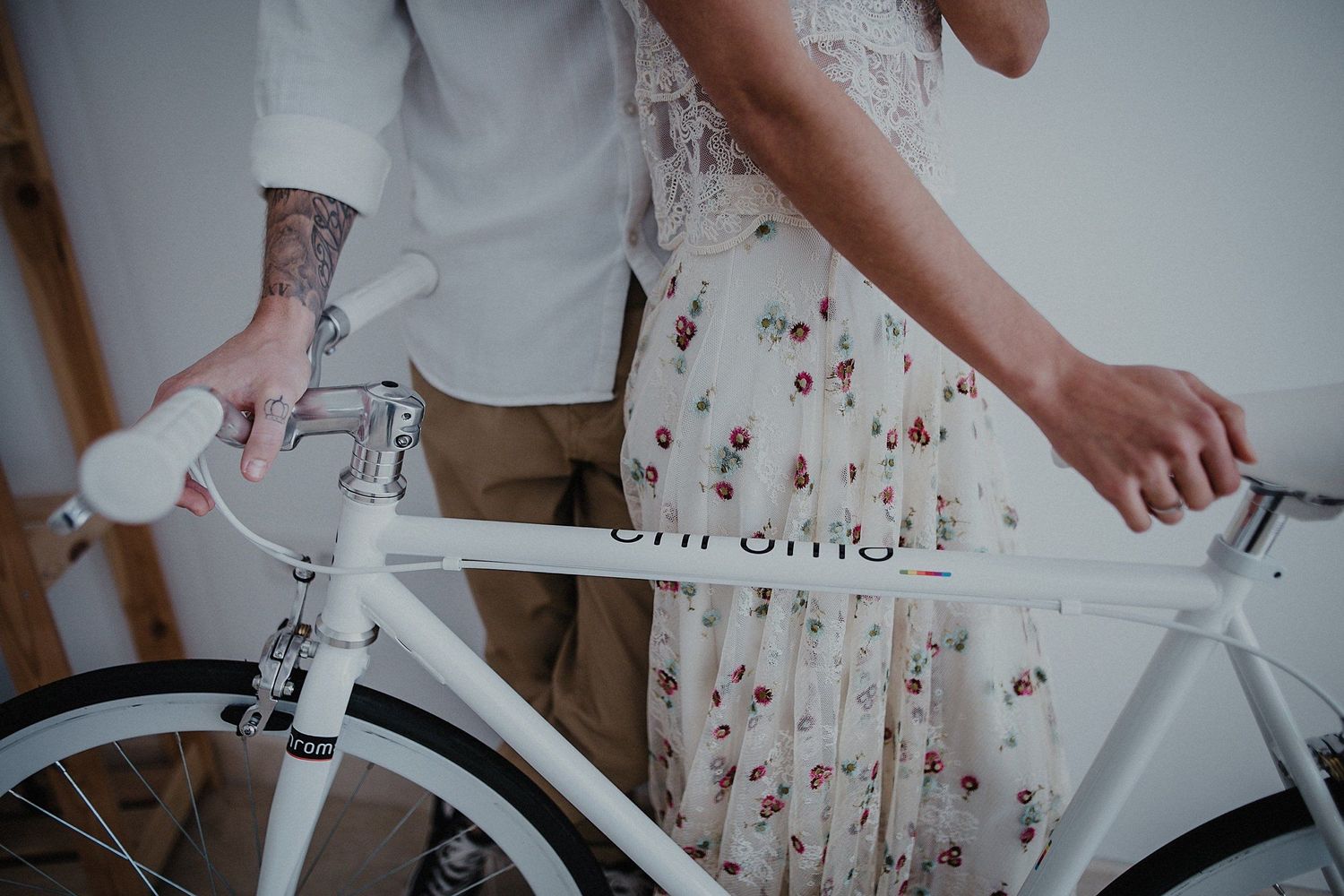 Close-up detail of tattooed hands and a floral dress against a white vintage bicycle.