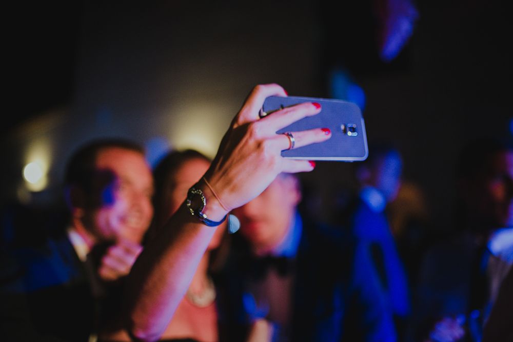 Hand holding up a phone to capture moments at a dimly lit event.