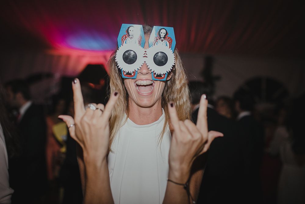 Party guest wearing decorative glasses making rock gesture at wedding reception.