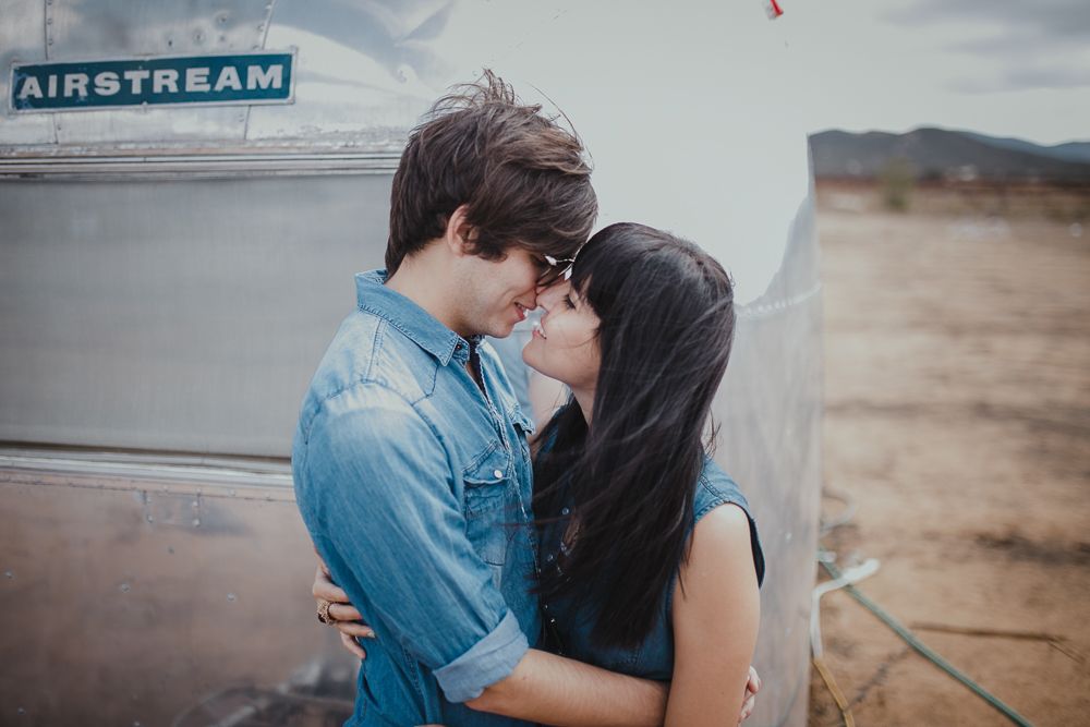 Romantic couple shares a close moment beside their Airstream trailer in a desert landscape.