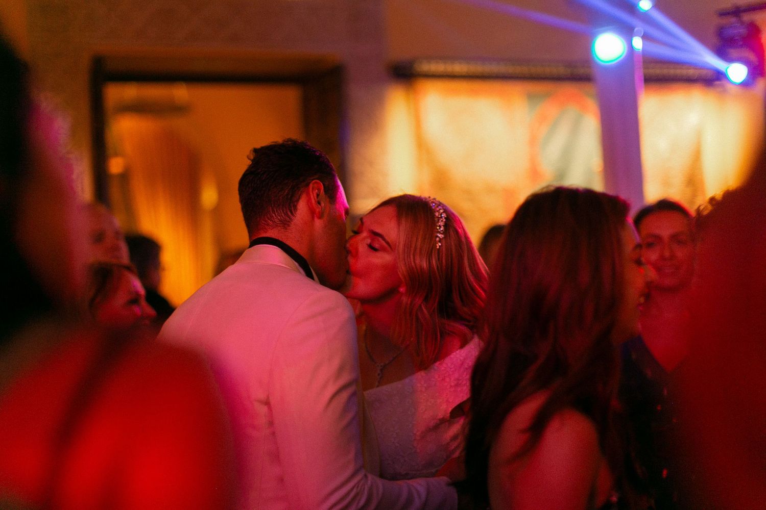 Couple sharing a romantic kiss on the dance floor at a wedding reception with colorful lights.