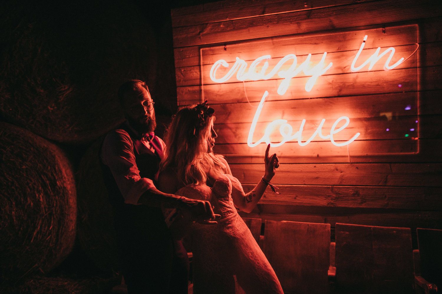 Couple poses beneath glowing neon 'Crazy in love' sign on rustic wood wall.