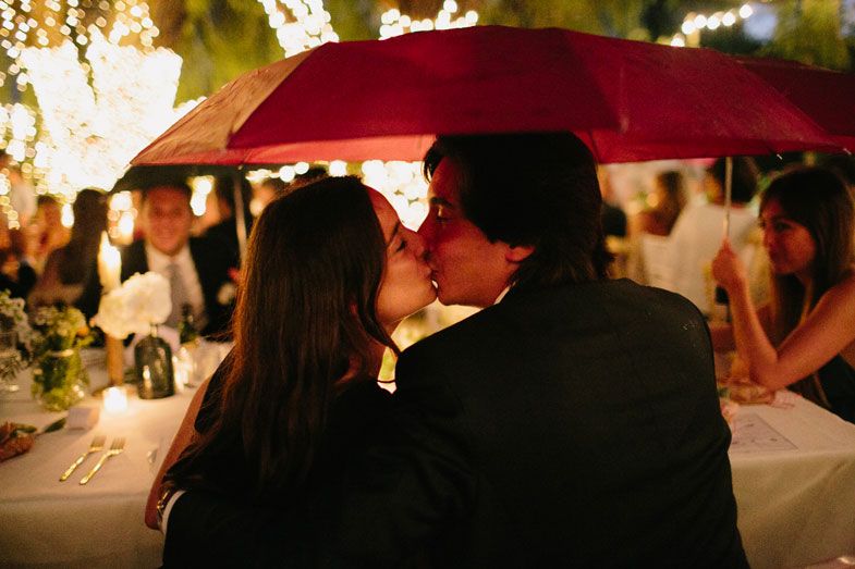 A romantic moment shared under a red umbrella during an evening wedding reception.
