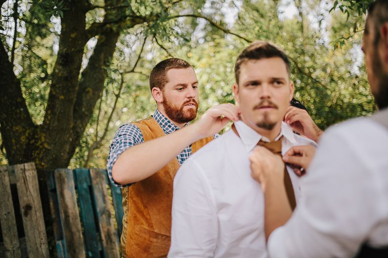 Wedding party members help adjust formal attire in outdoor garden setting.