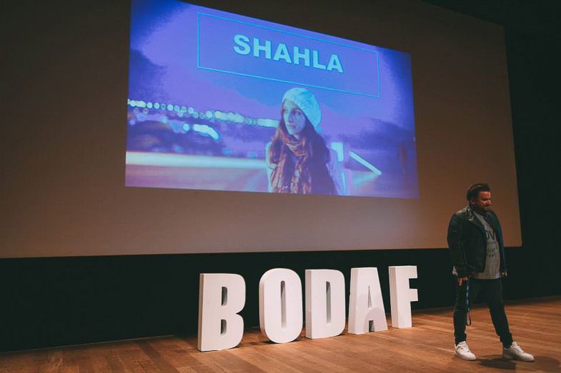 Stage presentation with large white letters spelling BODAE and projection screen showing text.