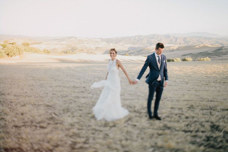 Couple walking hand in hand through rustic countryside field in wedding attire.