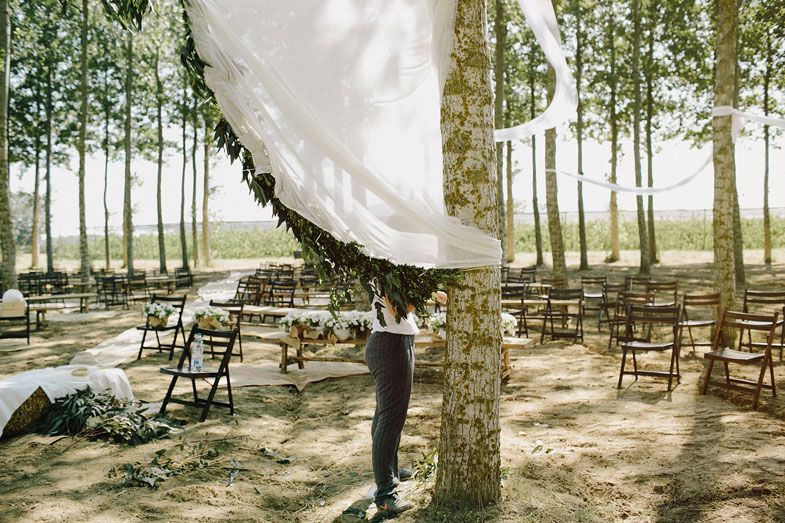 Person adjusting flowing white fabric drapes between trees at an outdoor event setup.