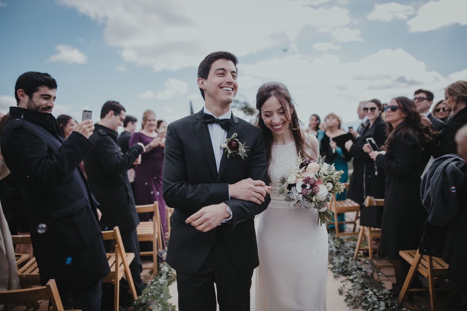 A joyful outdoor wedding ceremony with guests seated on wooden chairs in the background.