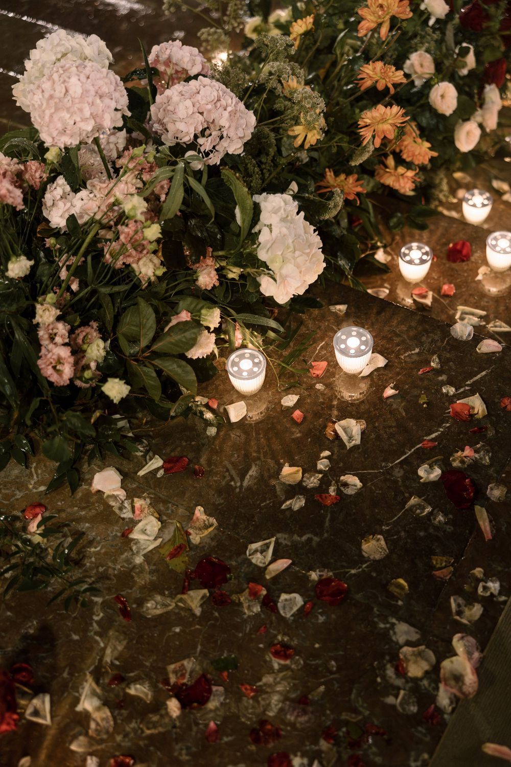 Romantic floral arrangement with candles and petals scattered on a dark surface.