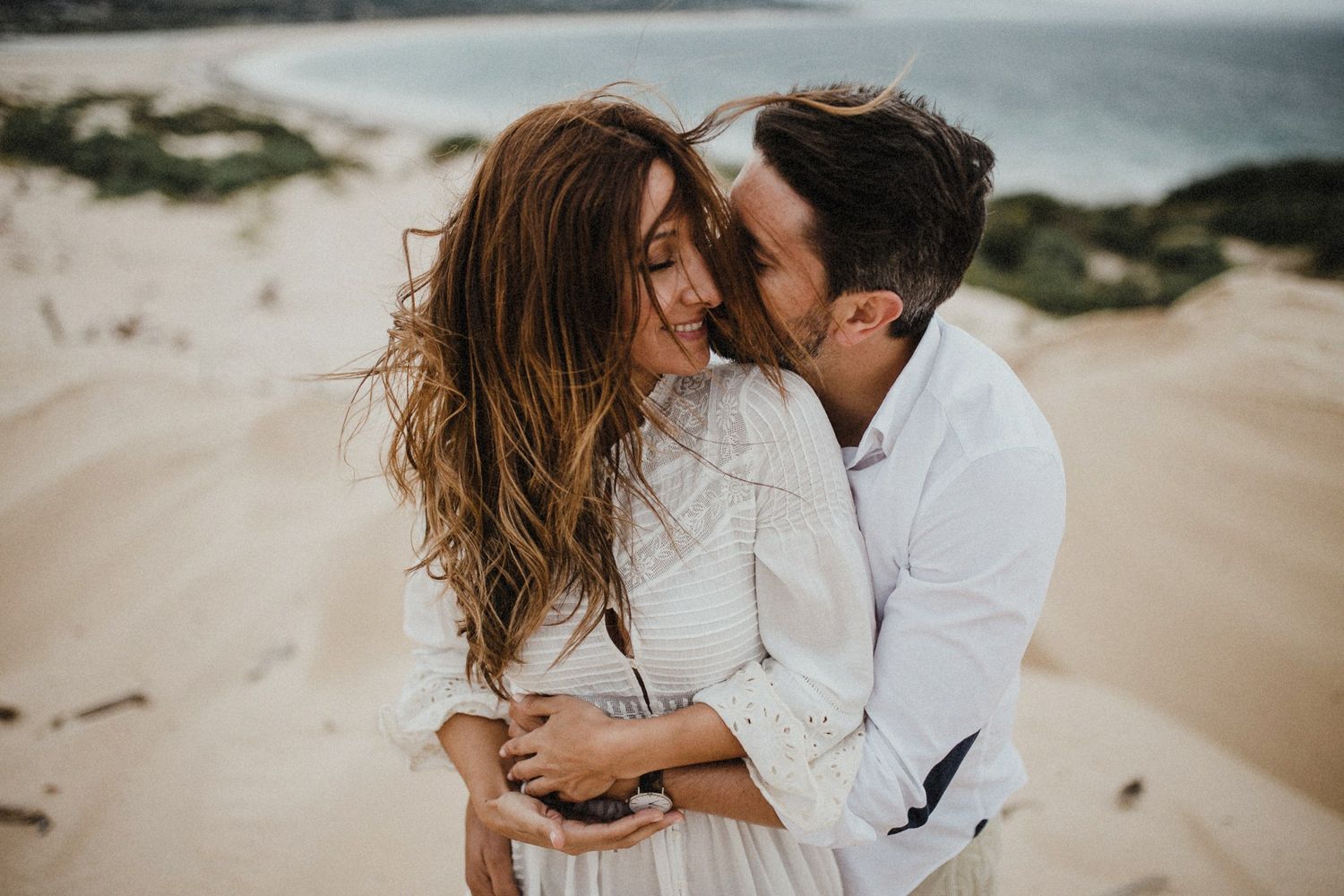 Intimate beach photo session with couple in white against coastal backdrop.