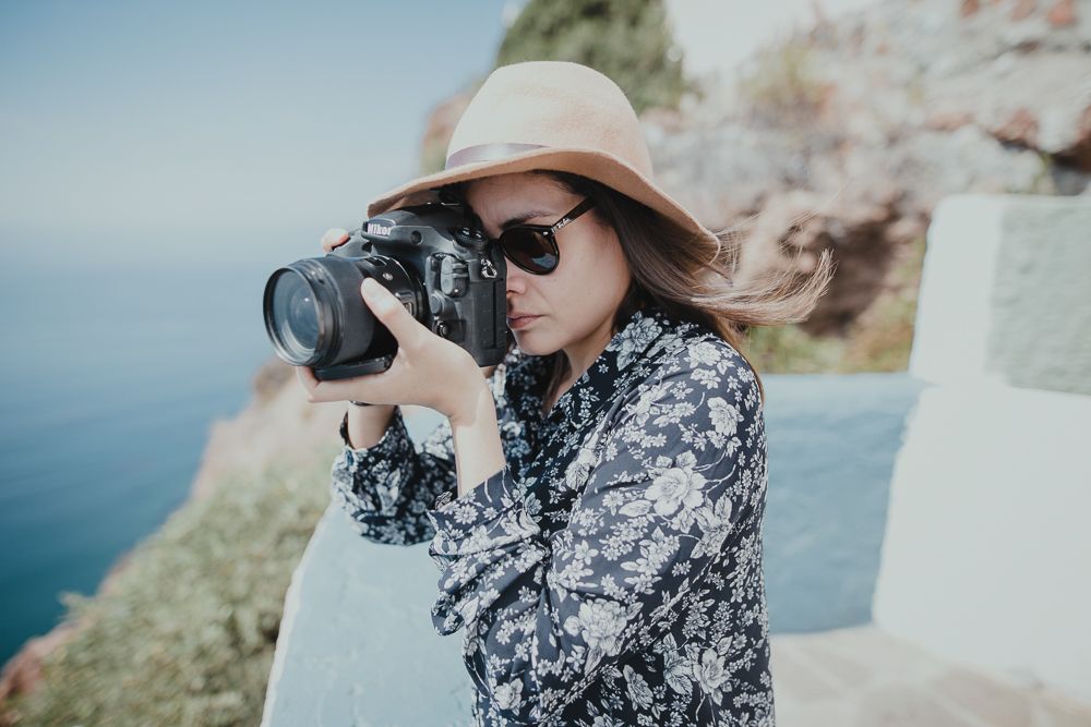 Photographer in floral dress and sun hat takes photos near coastal cliffs.