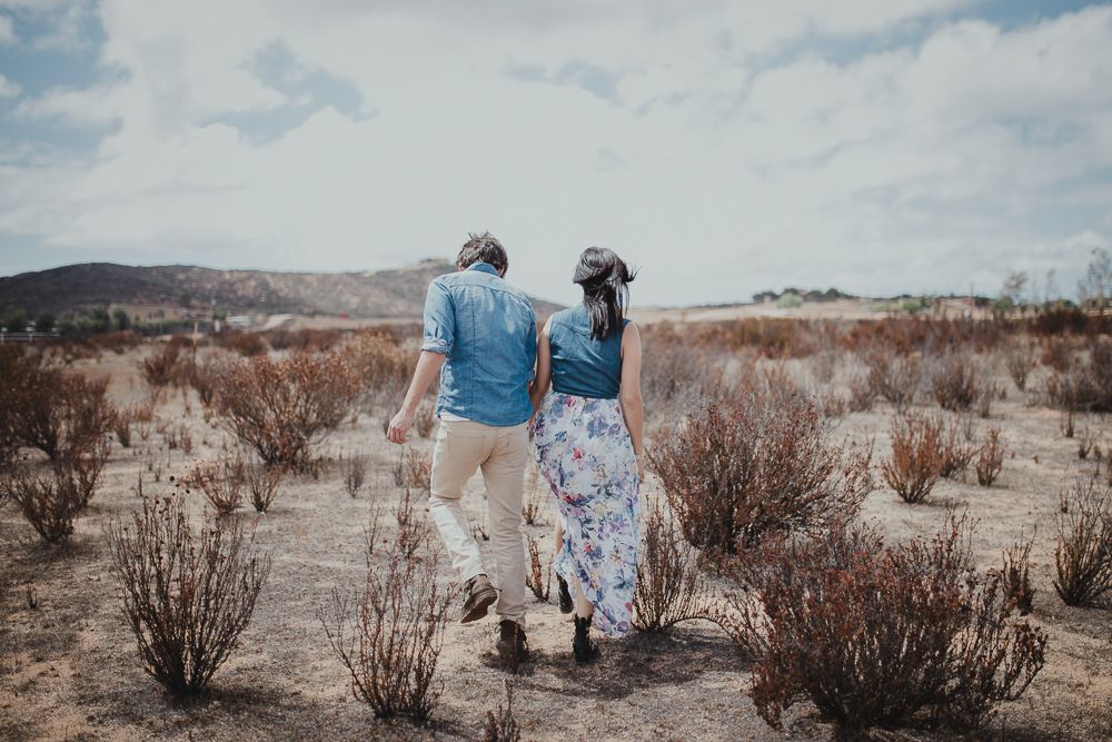 A couple walks hand in hand through desert brush towards distant mountains.