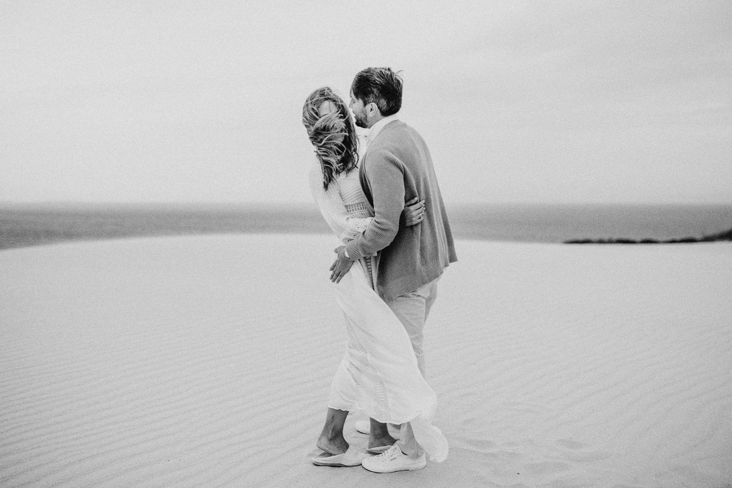 Black and white photo of couple dancing together on desert dunes.