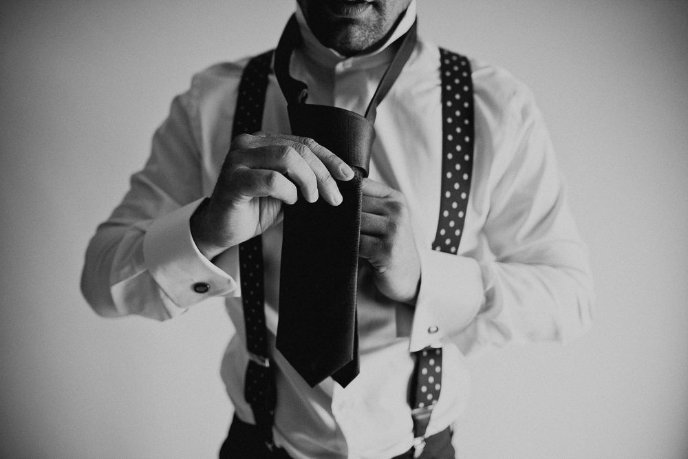 Hands adjusting black tie while wearing polka dot suspenders and white shirt.