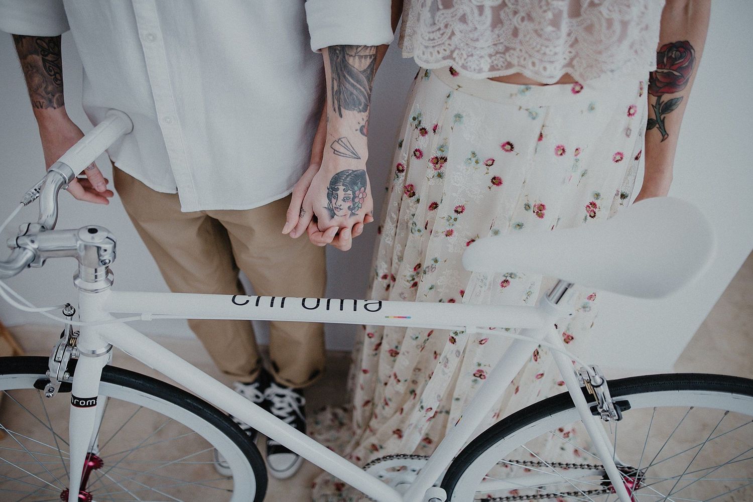 White vintage bicycle frames a stylish couple's lower bodies showing tattoos and floral dress details.