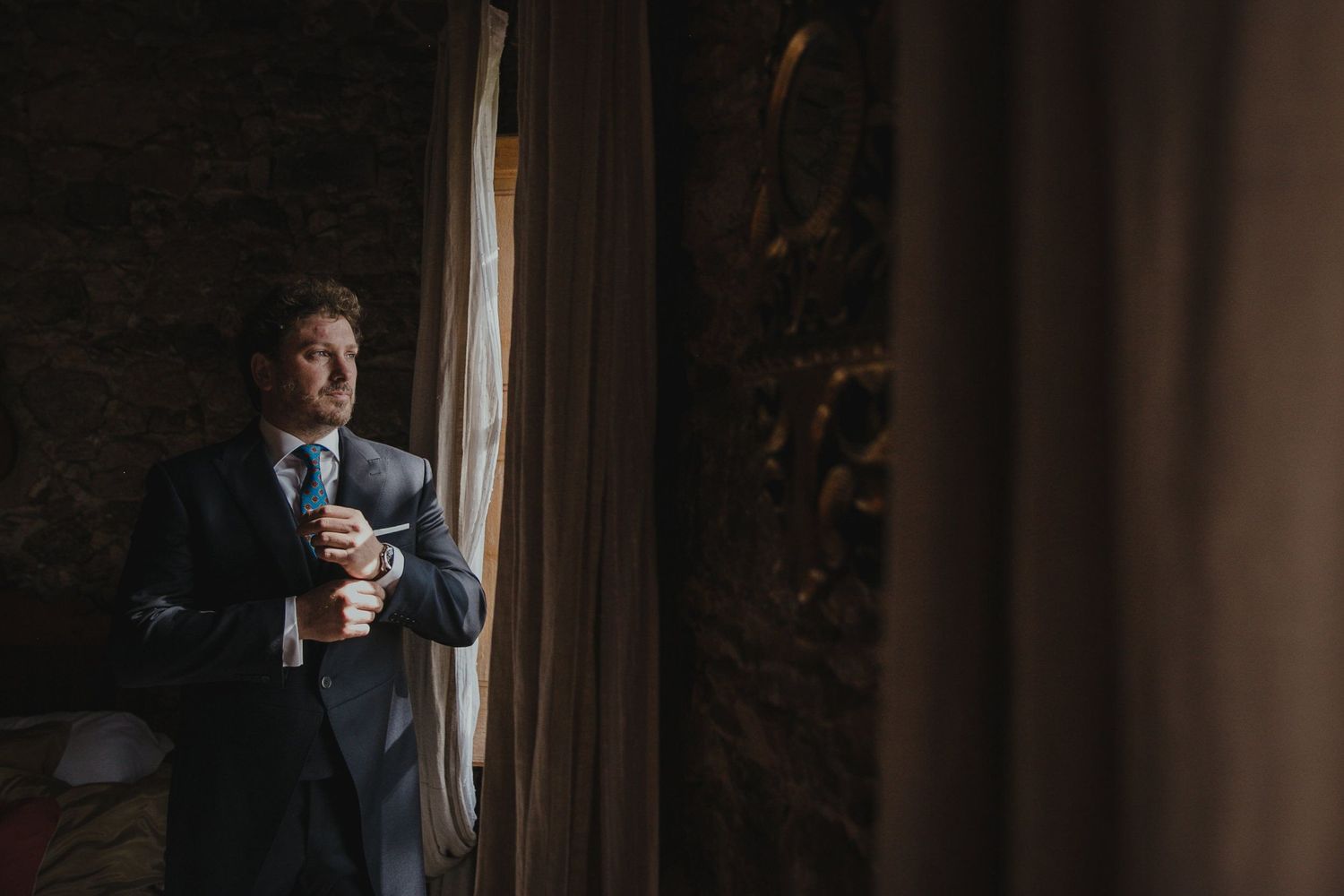 A person in a dark suit stands by a window in moody lighting.