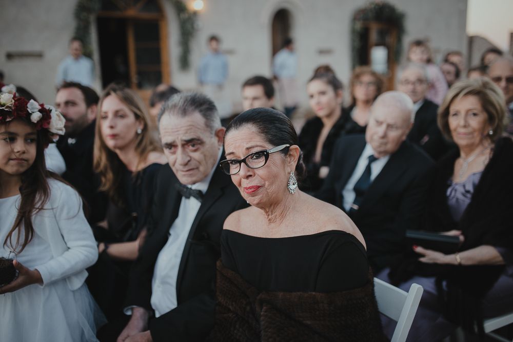 Wedding guests watch attentively during an outdoor ceremony beside a building with arched windows.