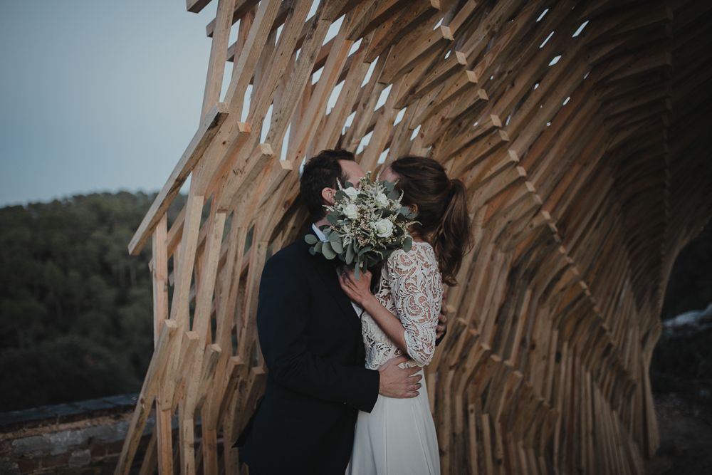 Newlyweds share an intimate moment under a wooden arch with mountain views.