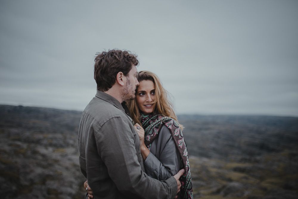 An intimate portrait of two people embracing on a dramatic hillside landscape.