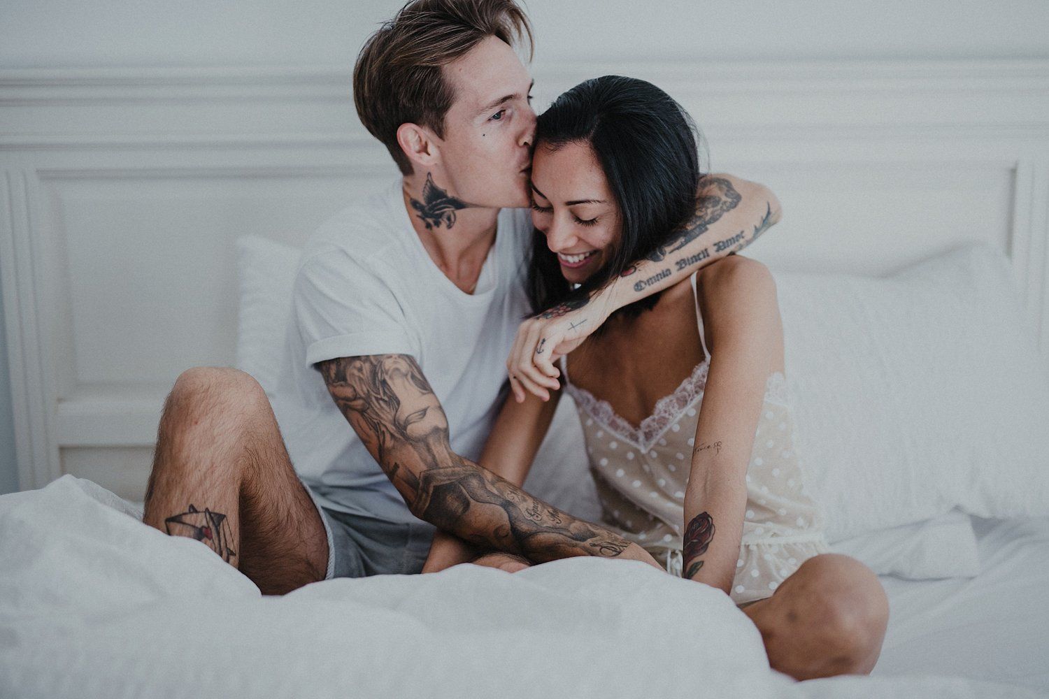 Couple embracing while sitting on bed with white linens and wall.