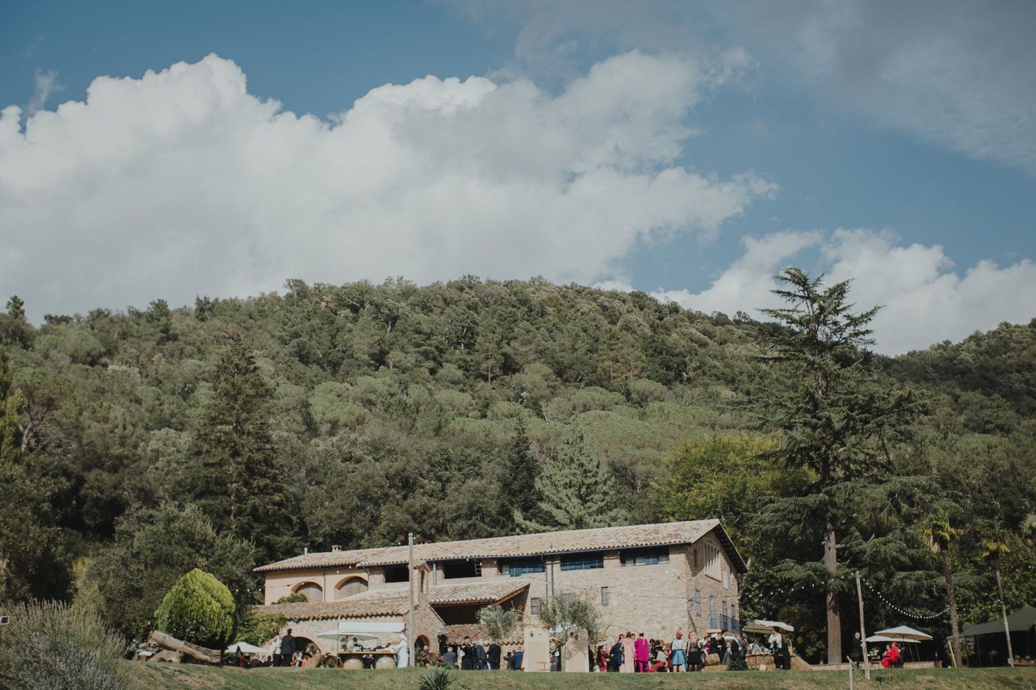 A rustic stone venue nestled in green hills with parked cars and guests outside.