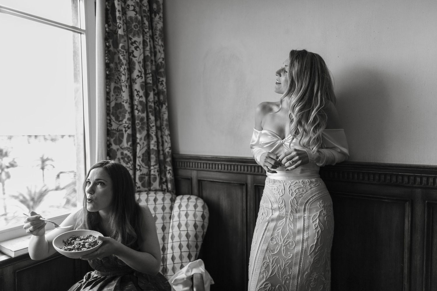 Black and white photo of two women in elegant dresses, one holding a bowl near a window, the other posing against a wall.