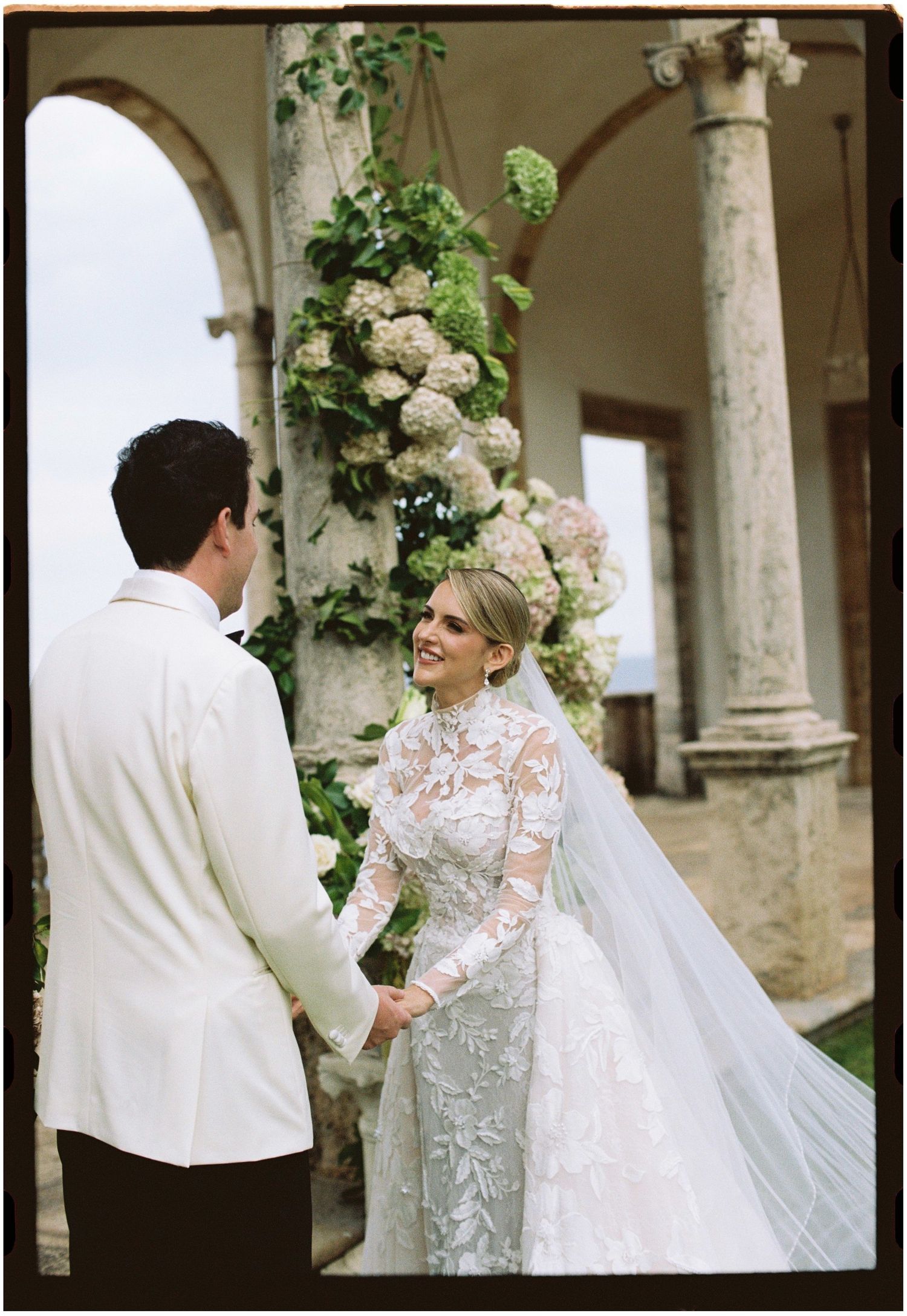 Bride in lace long-sleeve gown holds hands with groom in white tuxedo at elegant outdoor wedding ceremony with floral arch.