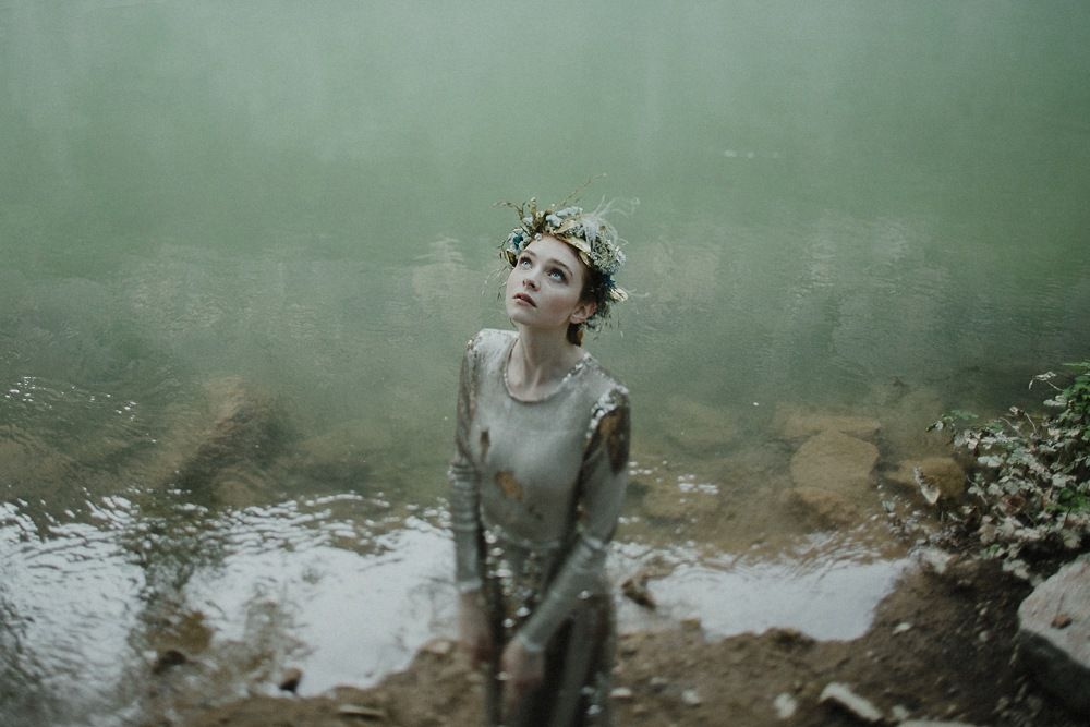 Figure in vintage dress and flower crown poses near misty water's edge.