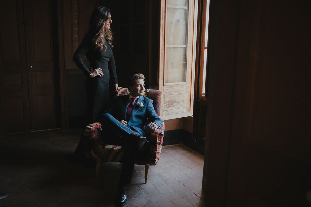 A couple poses in a moody, dark interior setting near a window.
