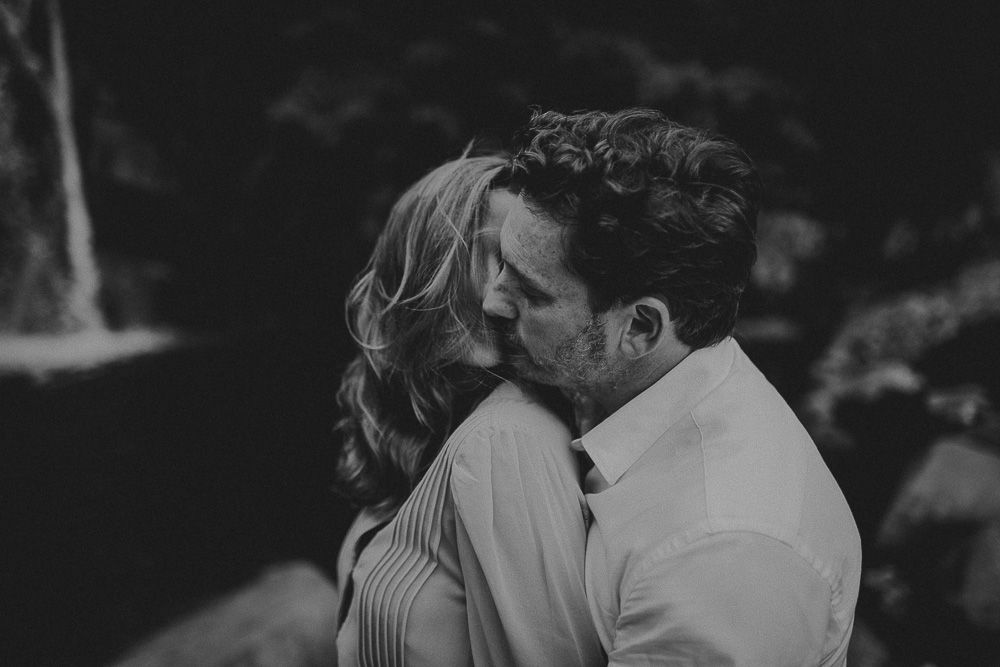 An intimate black and white portrait of a couple sharing a tender moment by a waterfall.