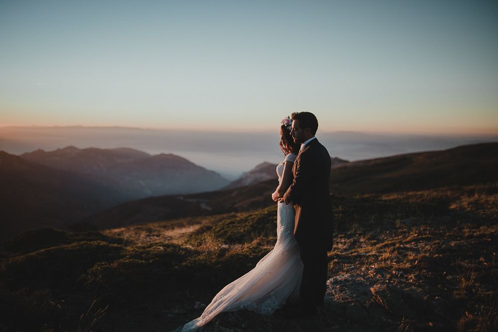 A wedding couple embraces while standing on a mountaintop during sunset.