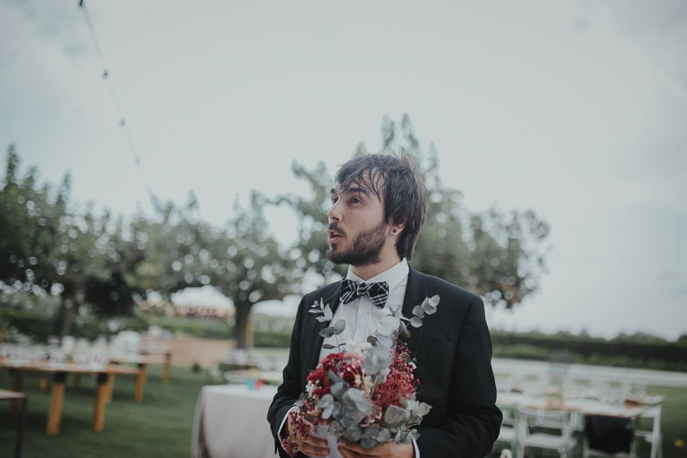 A groom in a black tuxedo with a pink boutonniere stands in an outdoor wedding venue with wooden tables.