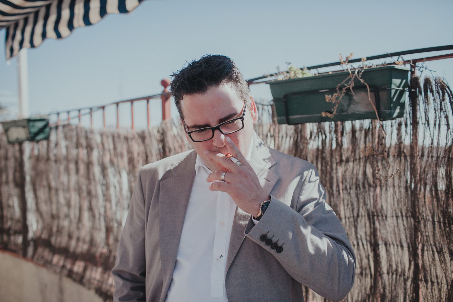 Person in grey suit and glasses smoking on a balcony with outdoor decor.
