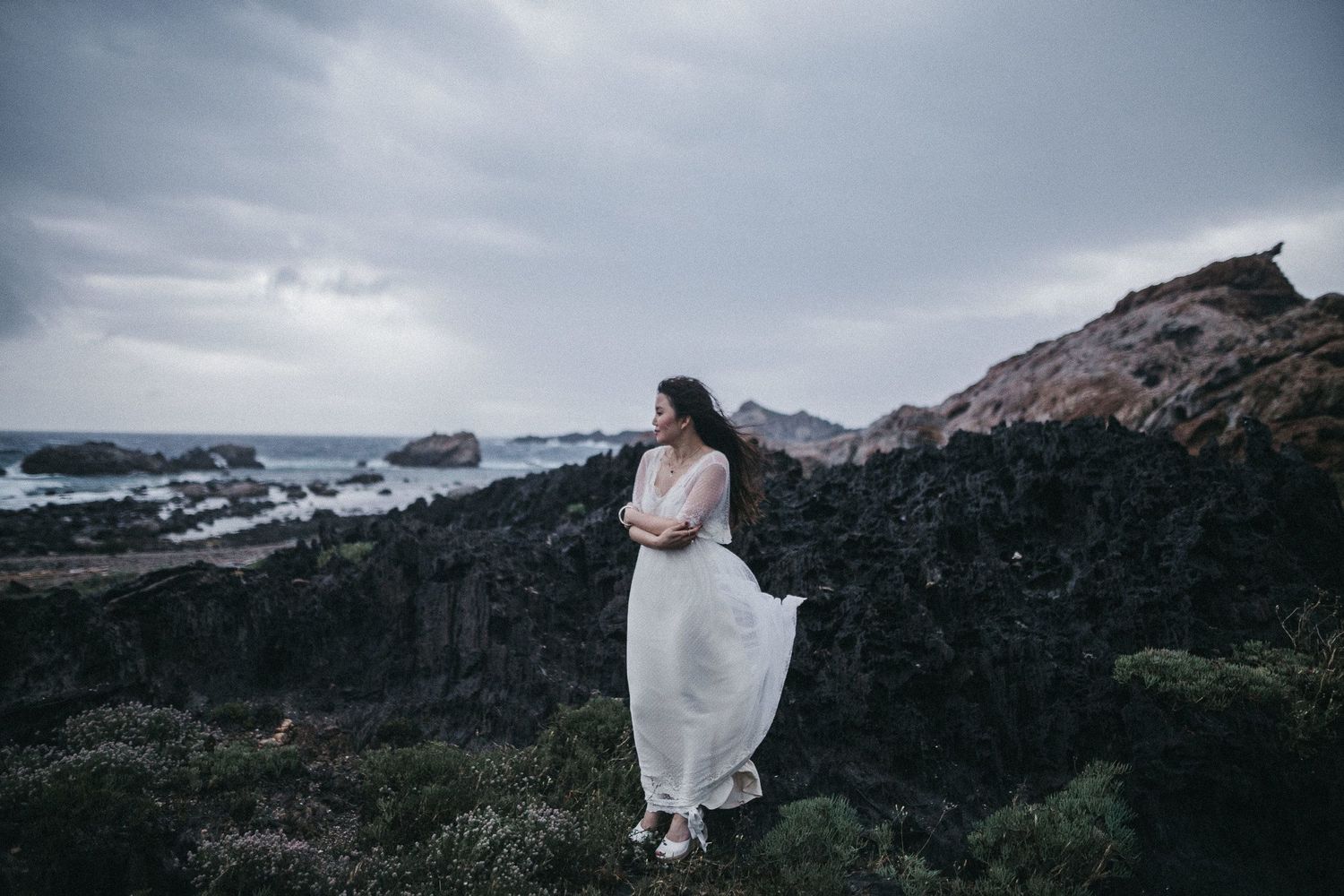 A flowing white dress catches the wind as a woman stands on dramatic coastal cliffs.