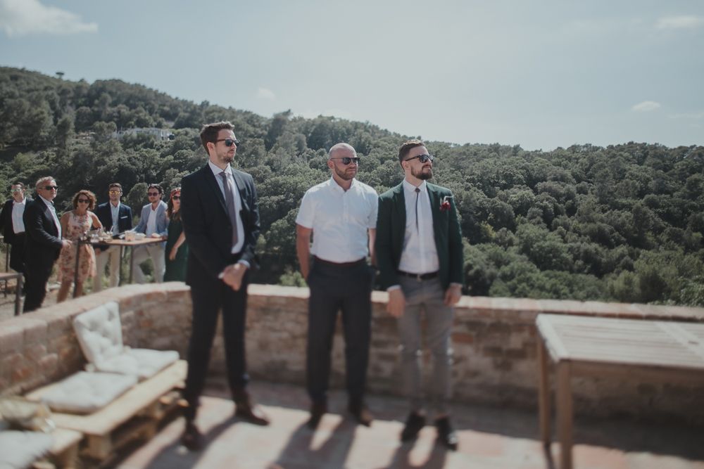 Three formally dressed individuals standing on a sunny terrace overlooking hills.