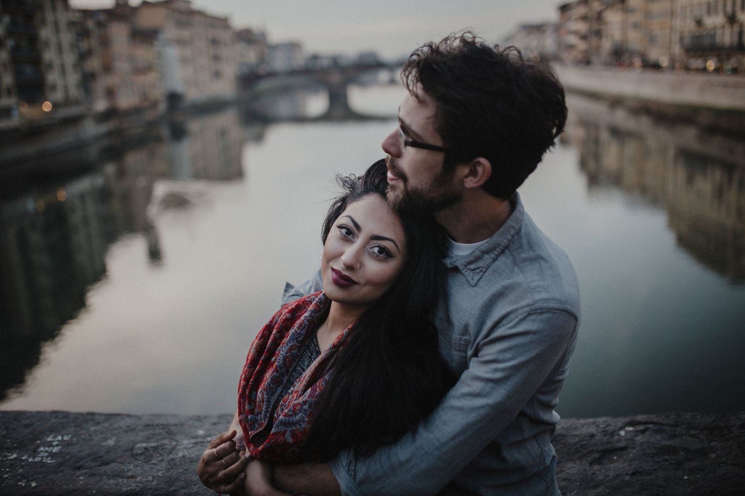 A romantic couple embraces on a bridge overlooking the Arno River in Florence during sunset.
