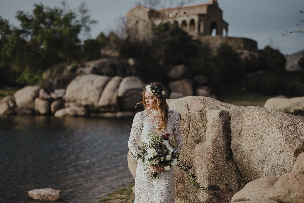 Lace-clad figure with bouquet poses against rocky lakeside landscape with stone building in background.