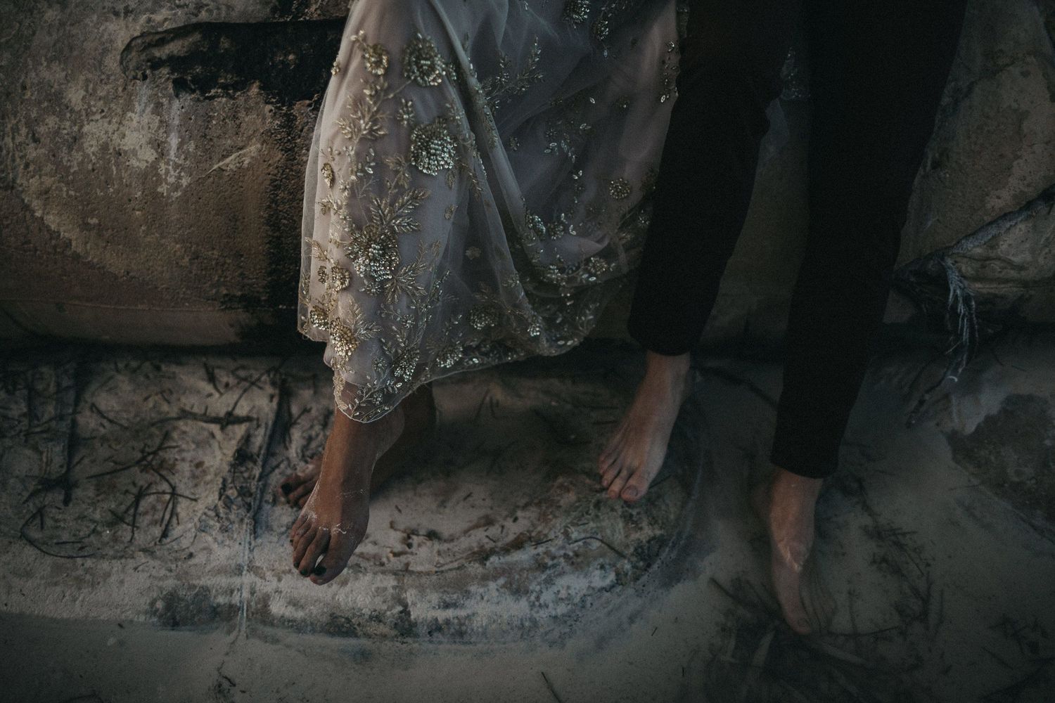 Close-up of bare feet and wedding dress hem against a weathered stone surface.