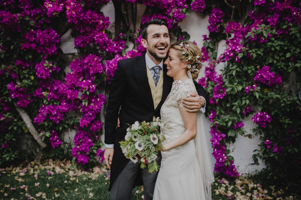 A joyful wedding couple laughs together in front of blooming purple bougainvillea flowers.