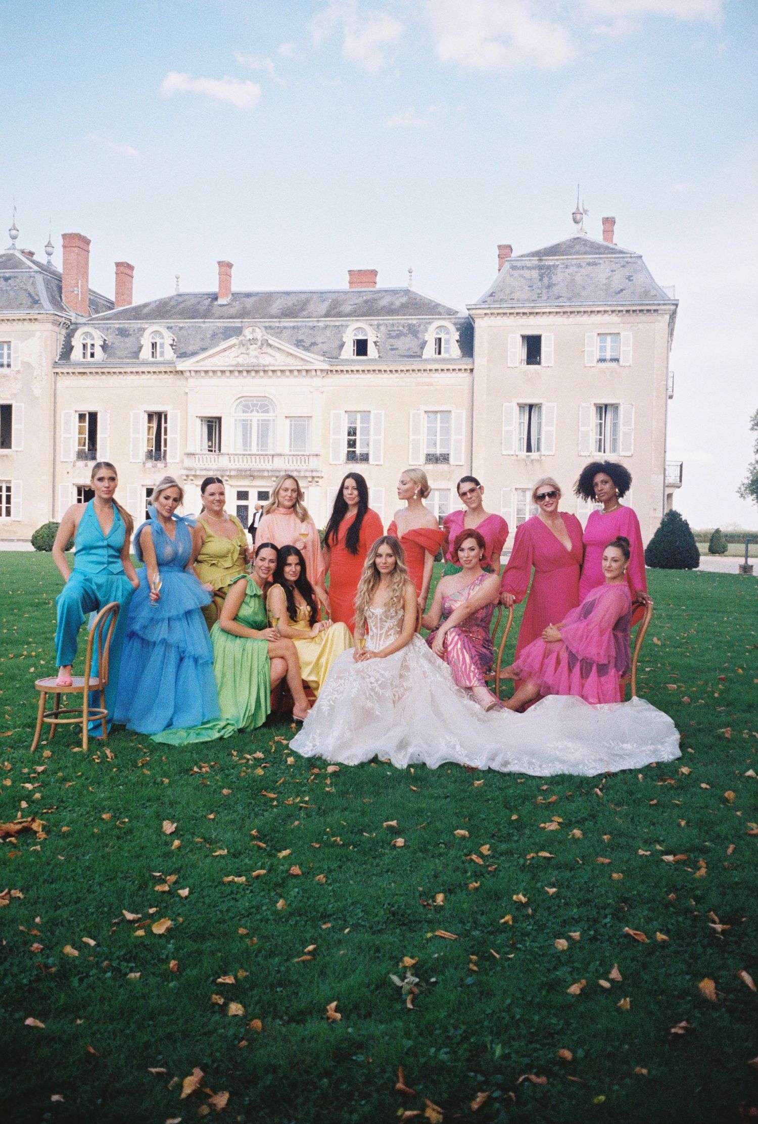 Bridal party in rainbow-colored dresses poses on lawn in front of elegant mansion.