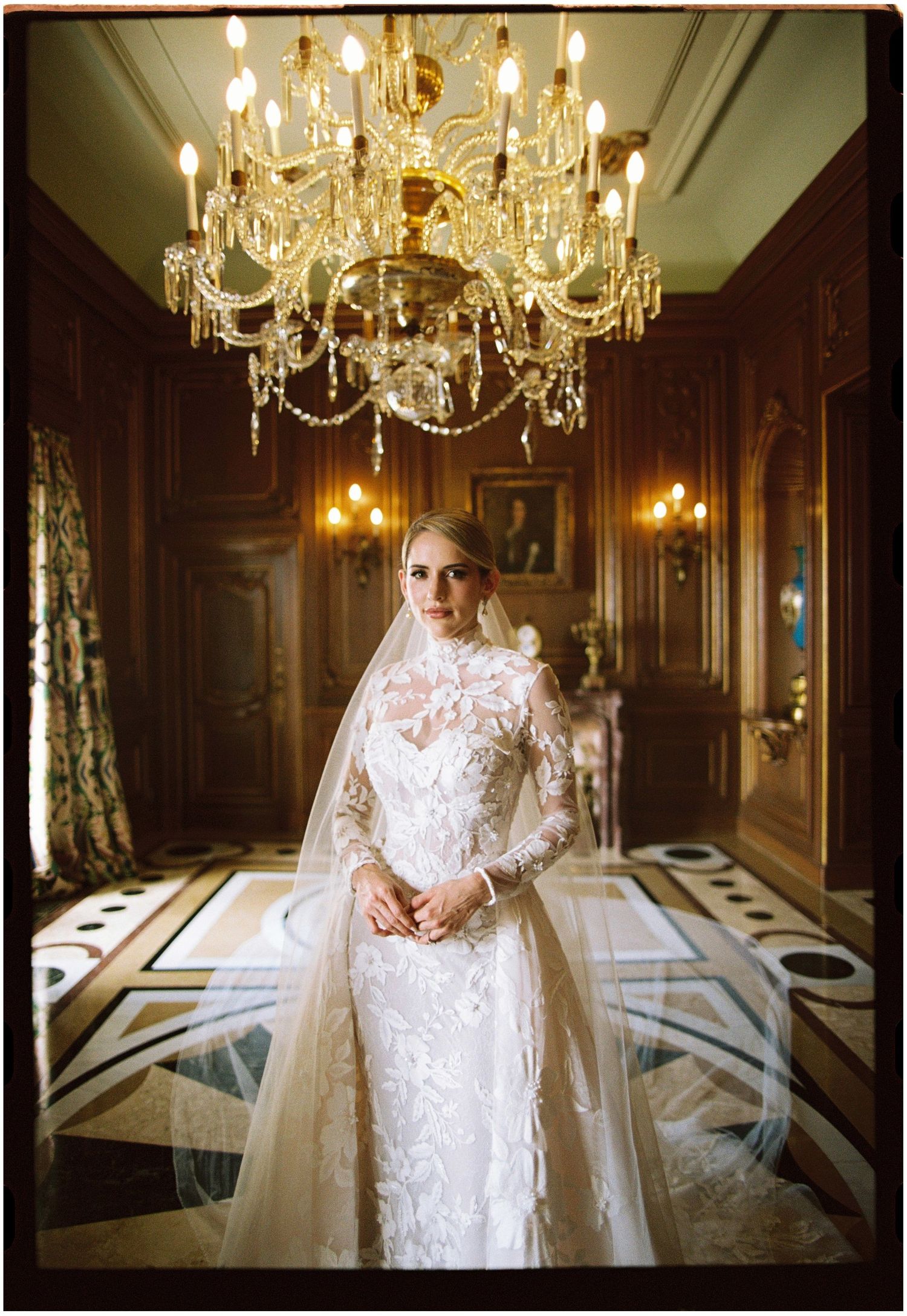 Bride in lace long-sleeve wedding gown with veil stands under crystal chandelier in elegant wood-paneled hall.