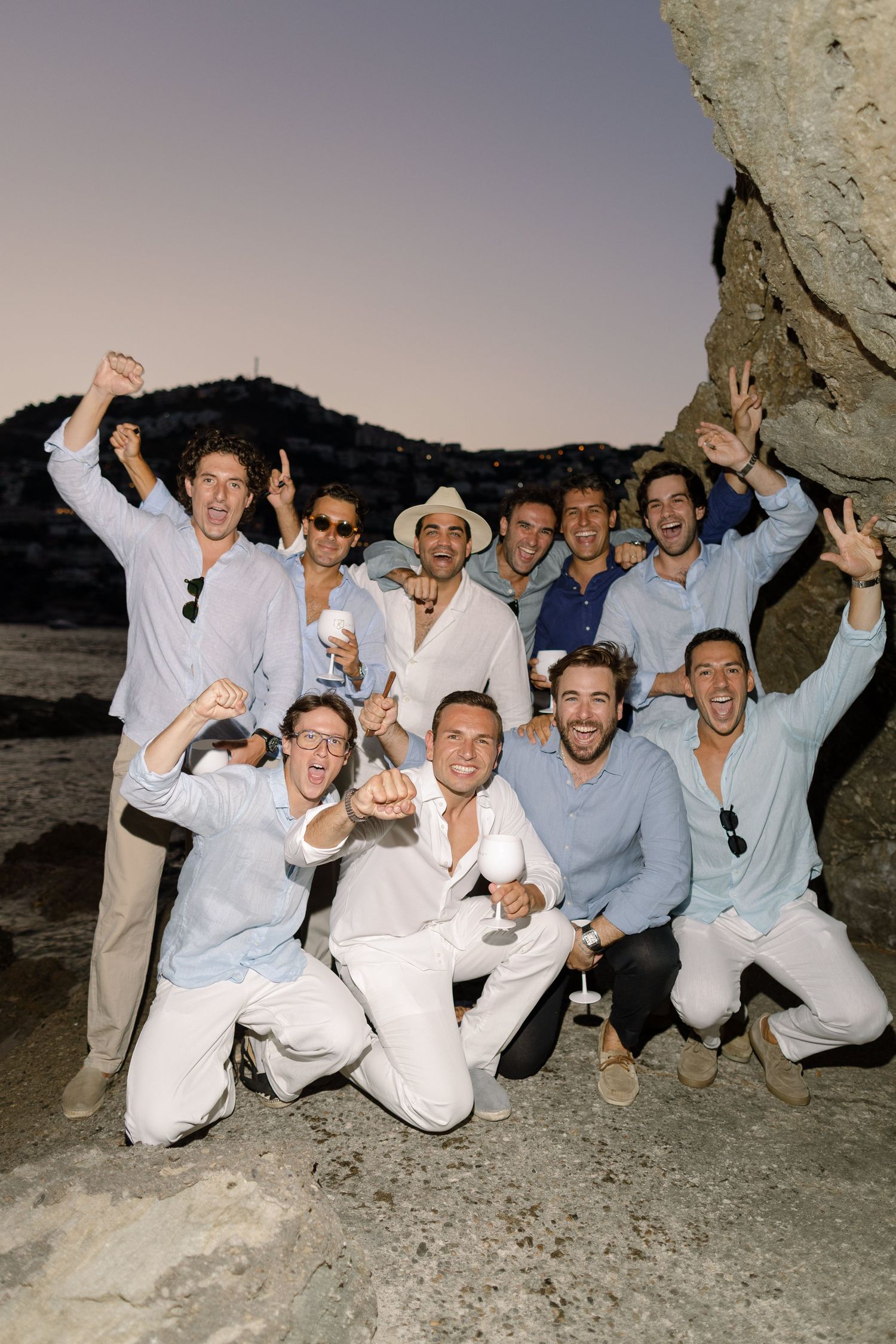 Group of men in white and light blue outfits celebrating outdoors on rocky terrain at dusk.