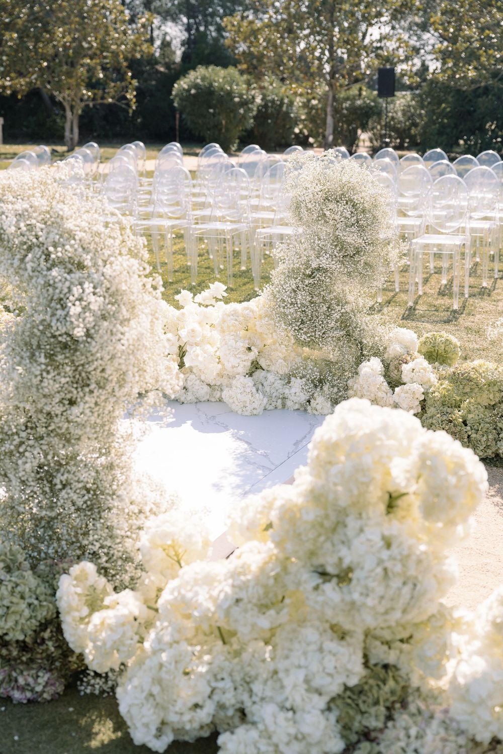 Outdoor wedding ceremony setup with white floral arrangements and rows of chairs among trees.