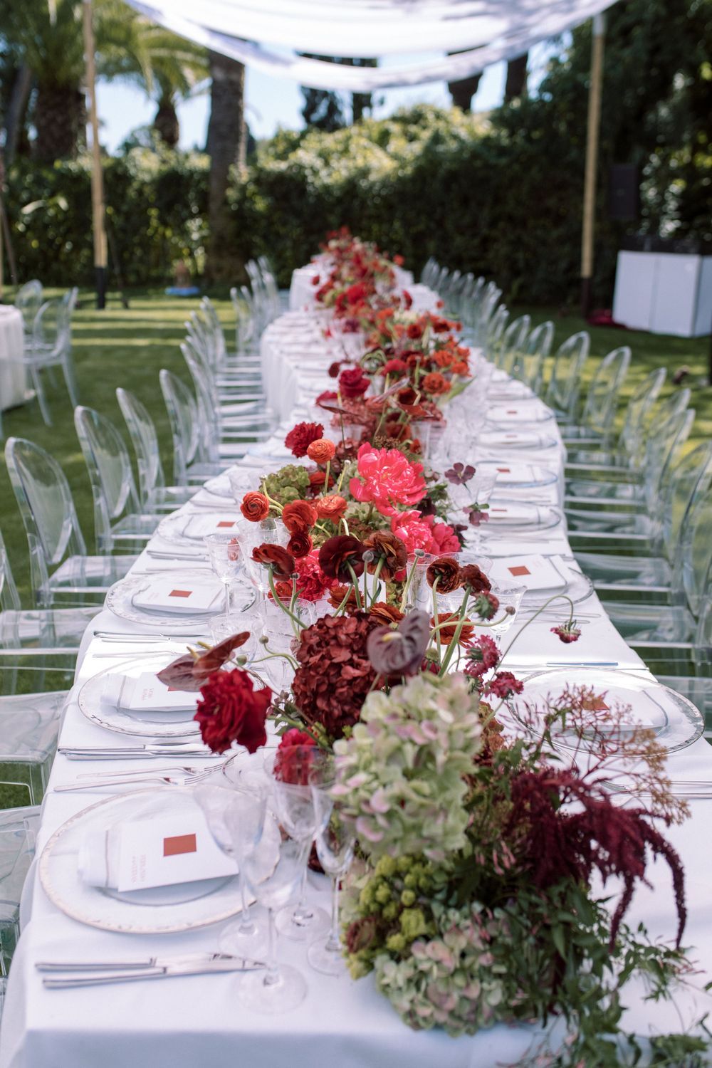 Long wedding reception table with white linens and lush burgundy floral centerpieces under outdoor canopy.