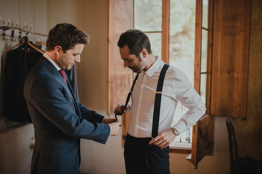 Two people helping each other with formal attire adjustments in a sunlit room.