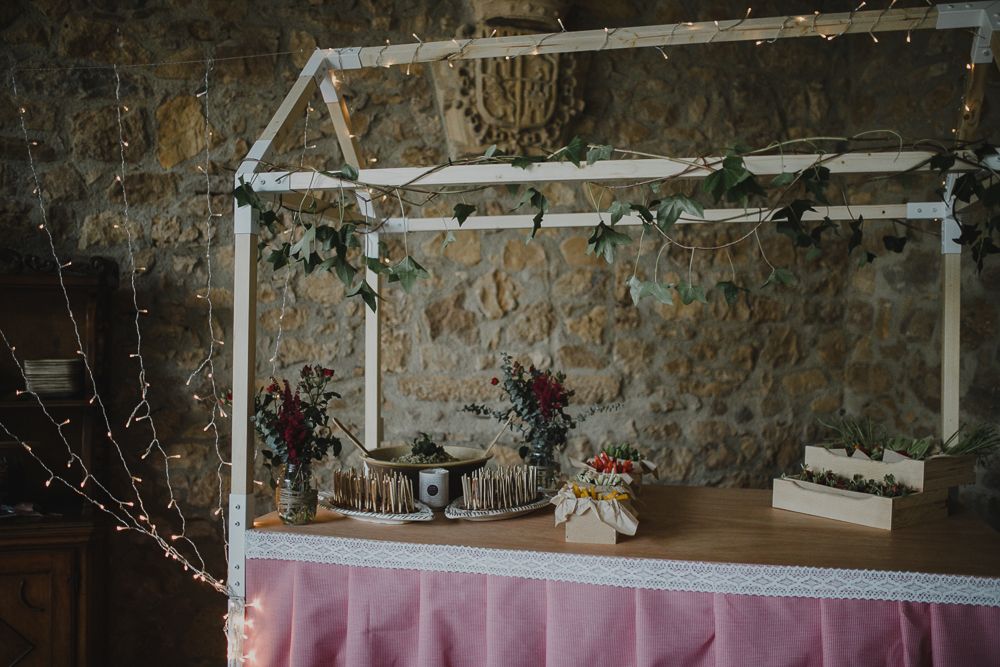 A rustic buffet table setup with white frame structure and pink tablecloth against a stone wall.