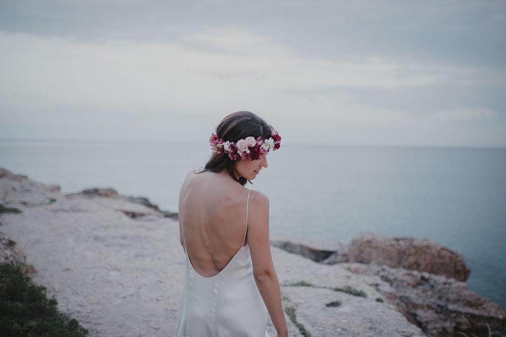 Elegant backless wedding dress shown against coastal backdrop as sun sets over ocean horizon.