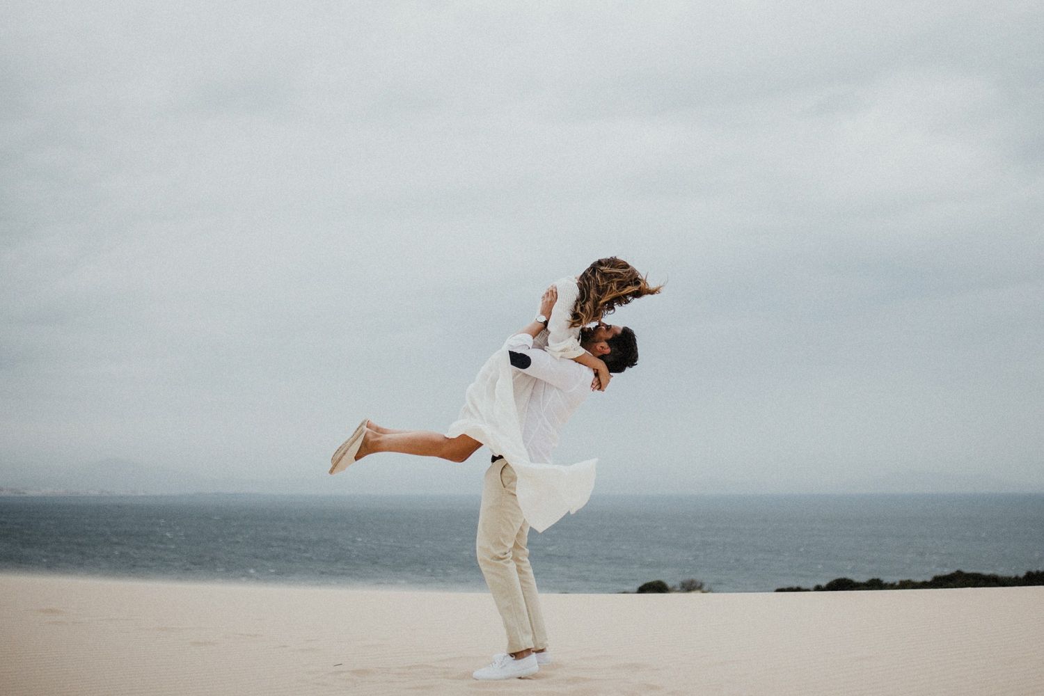 Joyful lift pose on beach with ocean and cloudy sky backdrop.