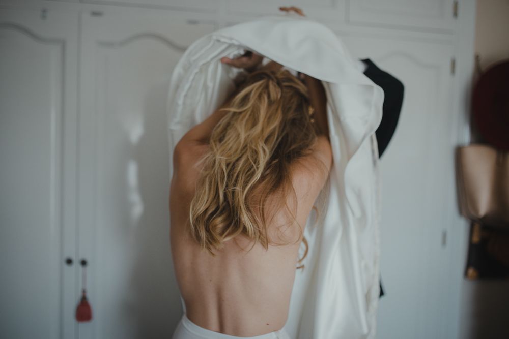 Back view of someone getting dressed in white wedding attire with blonde wavy hair.
