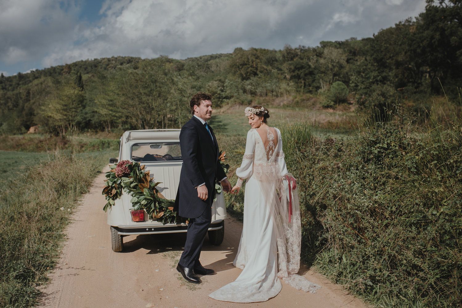 Newlyweds stand beside rustic truck on rural path surrounded by green hills.